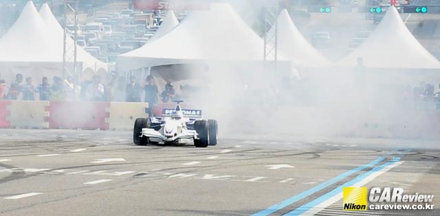 닉 하이드펠트의 F1 머신이 제자리에서 타이어를 태우며 선회하는 묘기(도우넛 주행)를 선보이며 연기를 뚫고 등장하고 있다.